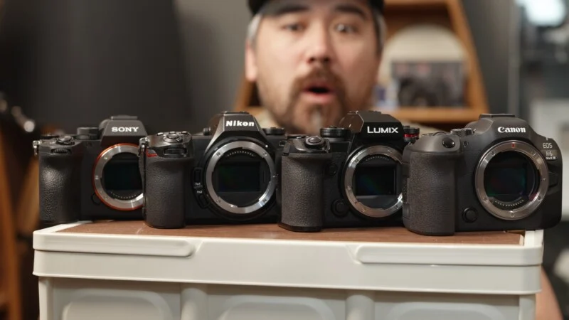 Un hombre sorprendido se encuentra detrás de cuatro cámaras sin espejo: Sony, Nikon, Lumix y Canon, alineadas sobre una superficie blanca.